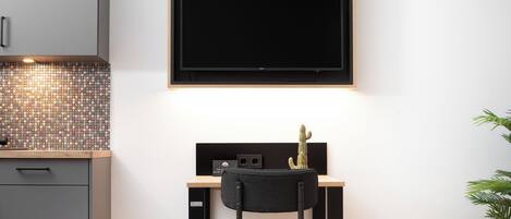 A modern kitchen with a tiled backsplash, a black-framed TV, a desk with a chair, and a cactus decoration.