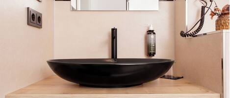 A modern bathroom with a black vessel sink, a mirror, a hairdryer, and a bottle of shampoo.