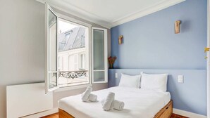 A bedroom with a large bed, a window with a view of buildings, and a blue wall. - SaintGermain Sojourn - Charming 2 bedroom in the Rive Gauche (Paris)