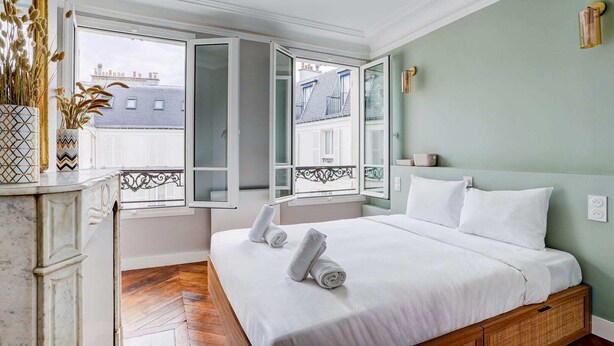 A bedroom with a bed, two open windows, a marble fireplace, and a view of buildings outside. - SaintGermain Sojourn - Charming 2 bedroom in the Rive Gauche (Paris)