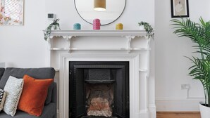 Una sala de estar con chimenea blanca, un sofá gris con cojines naranja y un espejo redondo sobre la repisa.