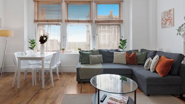 Una sala de estar moderna con un sofá gris, sillas blancas, una mesa de centro de vidrio y una mesa de comedor con un jarrón de flores.