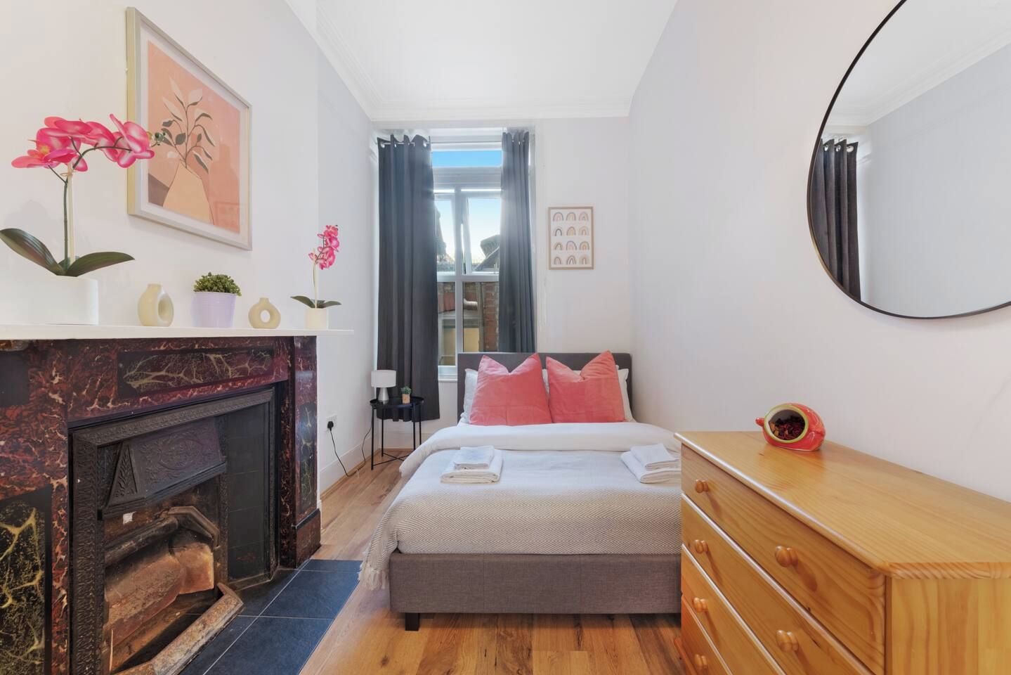 A bedroom with a fireplace, a bed with pink pillows, a wooden dresser, and a mirror on the wall.