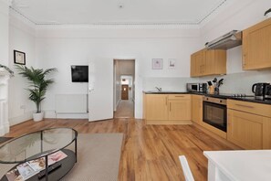 A modern kitchen with wooden cabinets, a microwave, and a stove. There is a glass table with magazines and a remote control. - Athenaeum Pl N10 | Muswell Hill | First Floor 1BD (London)