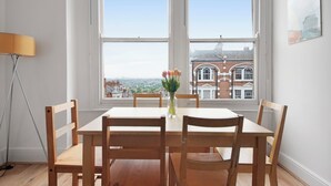 A dining area with a table and chairs, a vase with flowers, and a view of buildings through large windows.