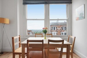 Ein Essbereich mit Tisch und Stühlen, einer Blumenvase und Blick auf Gebäude durch große Fenster.