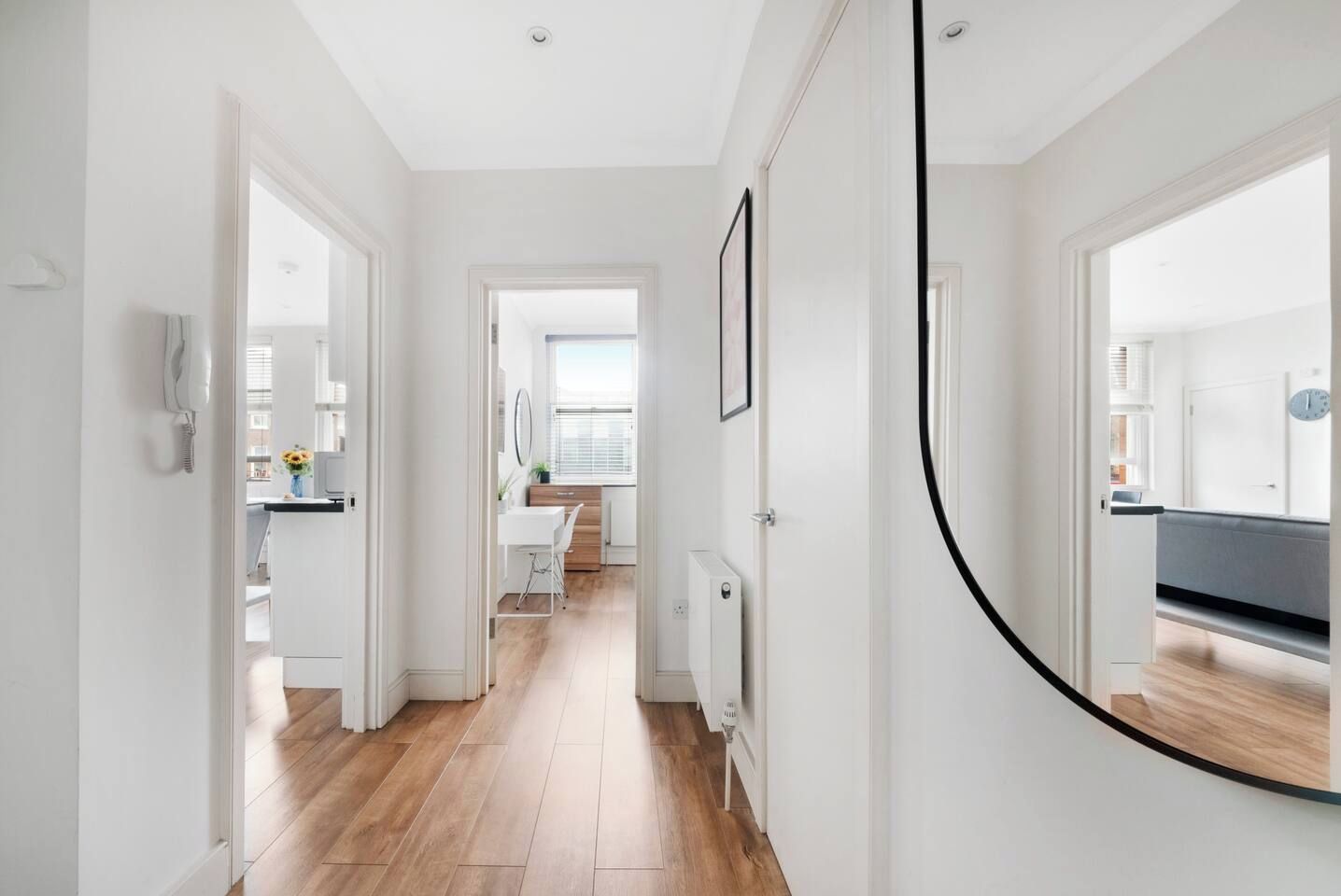 A hallway with a wooden floor, white walls, and a door leading to a room with a bed and a window.