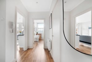 A hallway with a wooden floor, white walls, and a door leading to a room with a bed and a window.