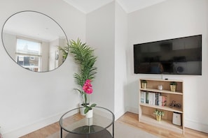 A modern living room with a round glass table, a large mirror, a potted plant, a bookshelf, and a flat-screen TV.