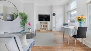 Ein modernes Wohnzimmer mit einer Couch, einem Couchtisch, einem Fernseher, einem Bücherregal und einem Essbereich mit Tisch und Stühlen.