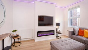 A modern living room with a flat-screen TV mounted on the wall, a fireplace below, a grey sofa with orange cushions, and a small round table with a plant.