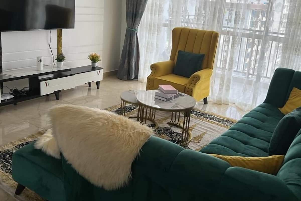Ein Wohnzimmer mit an der Wand montiertem Fernseher, einem gelben Sessel, einem grünen Sofa und einer Deckenleuchte.