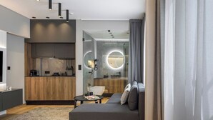 A modern hotel room with a sofa, a small table, a kitchen area, and a bathroom with a glass partition. - Aristotelian Virtues - Enkrateia (Thessaloniki)
