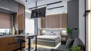 Quarto de hotel moderno com cozinha compacta, cama com roupa de cama cinzenta, sofá cinza e TV fixada na parede.