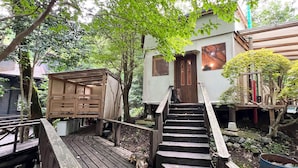 Eine Holzterrasse führt zu einem Haus mit Treppe, umgeben von Bäumen und Grünpflanzen.