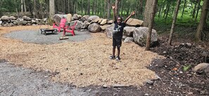 A forested area with a pile of wood chips, a red chair, and a metal fire pit.