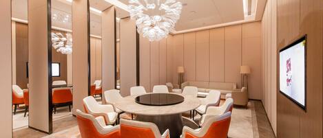 A modern conference room with a round table, orange chairs, a large chandelier, and a screen on the wall.