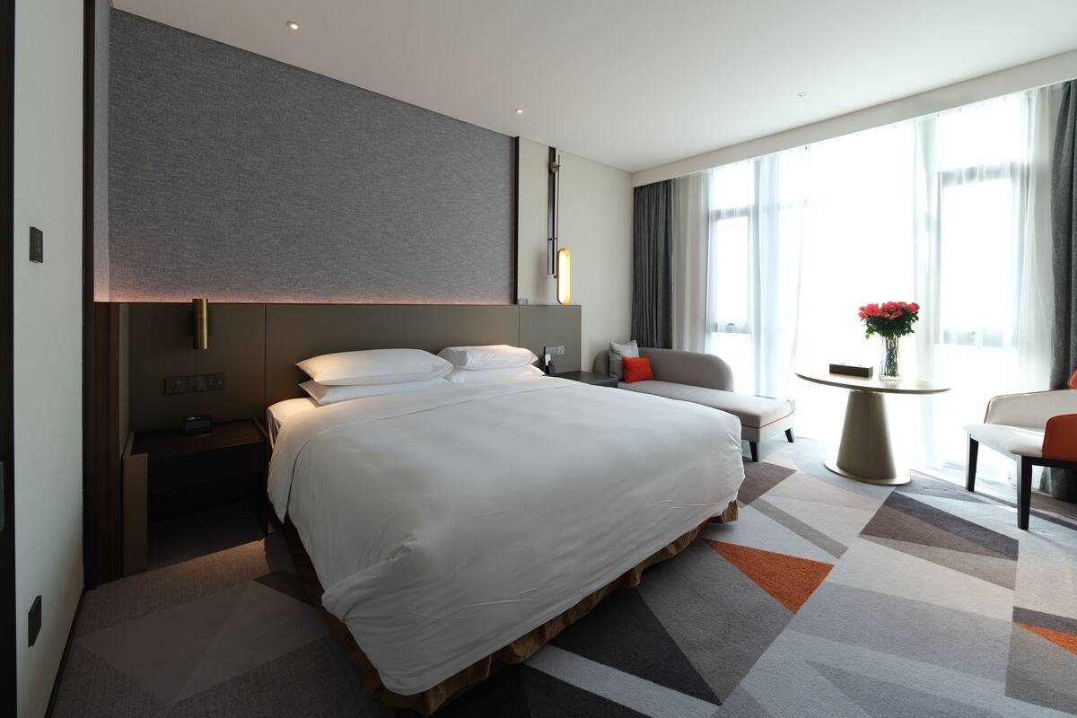 A modern hotel room with a large bed, a bedside table, a small desk, a sofa, a round table, and a view of the city through large windows.