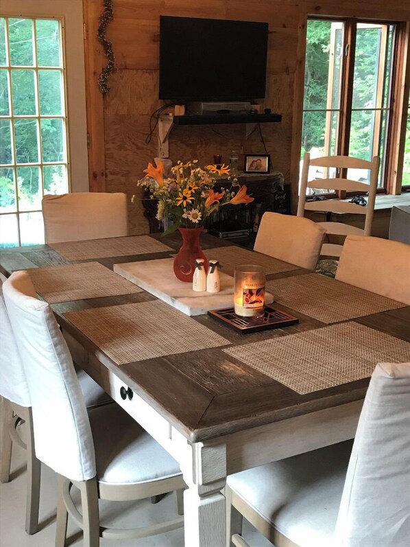 Una mesa de comedor de madera con individuales, sillas blancas acolchadas, un jarrón con flores, una vela y un televisor montado en la pared.