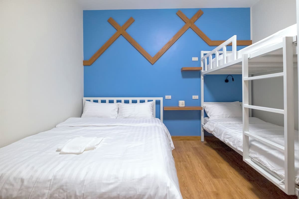 A room with two beds, one with a white headboard and the other with a white bunk bed. There is a blue wall with wooden X-shaped decorations and a small shelf with a lamp.