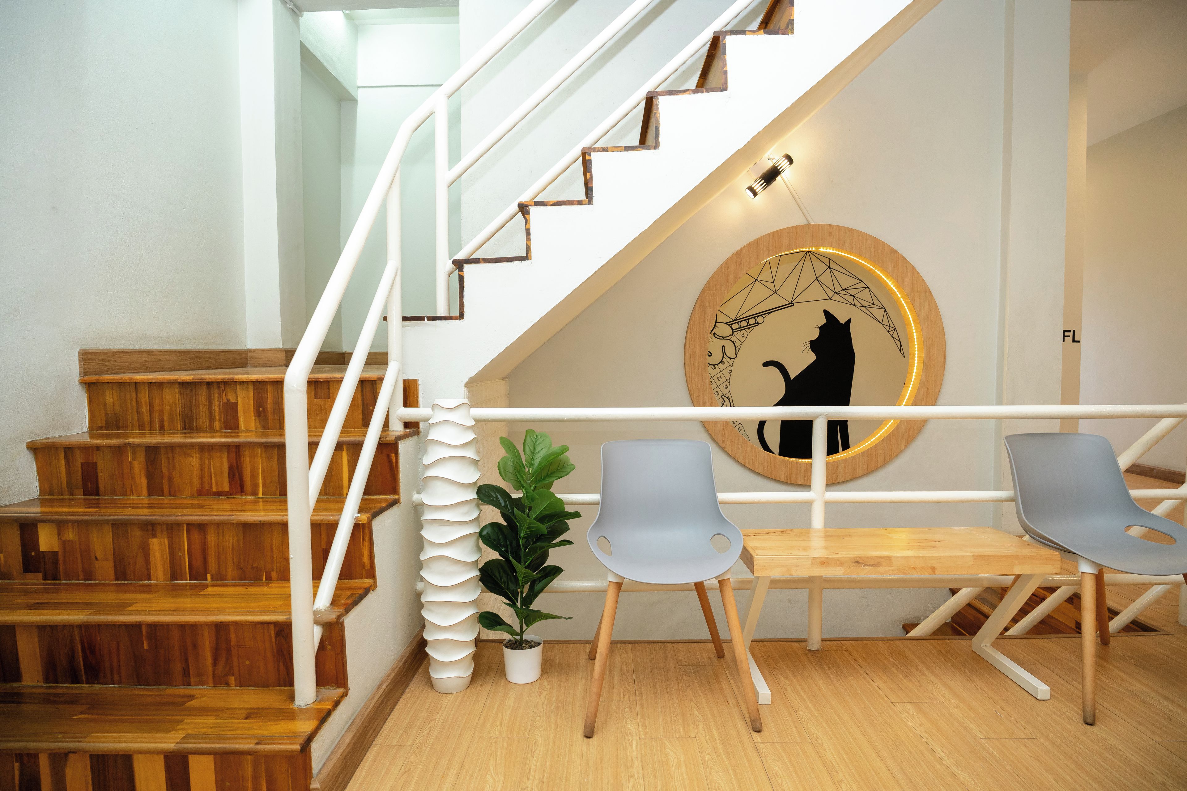 Una escalera con peldaños de madera y barandillas blancas, un adorno circular en la pared con silueta de un gato, y dos sillas grises con una pequeña planta en maceta a su lado.