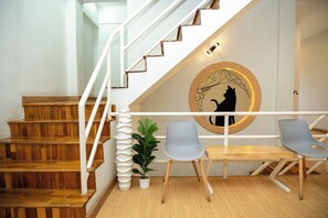 Una escalera con peldaños de madera y barandillas blancas, un adorno circular en la pared con silueta de un gato, y dos sillas grises con una pequeña planta en maceta a su lado.