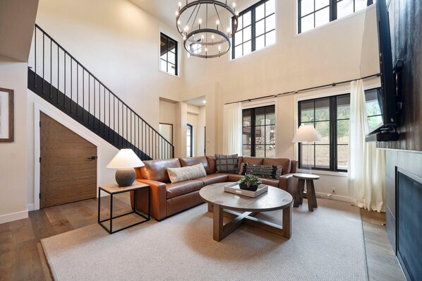 A spacious living room with a large sectional sofa, a coffee table, and a TV. A staircase with a black railing leads to an upper level.