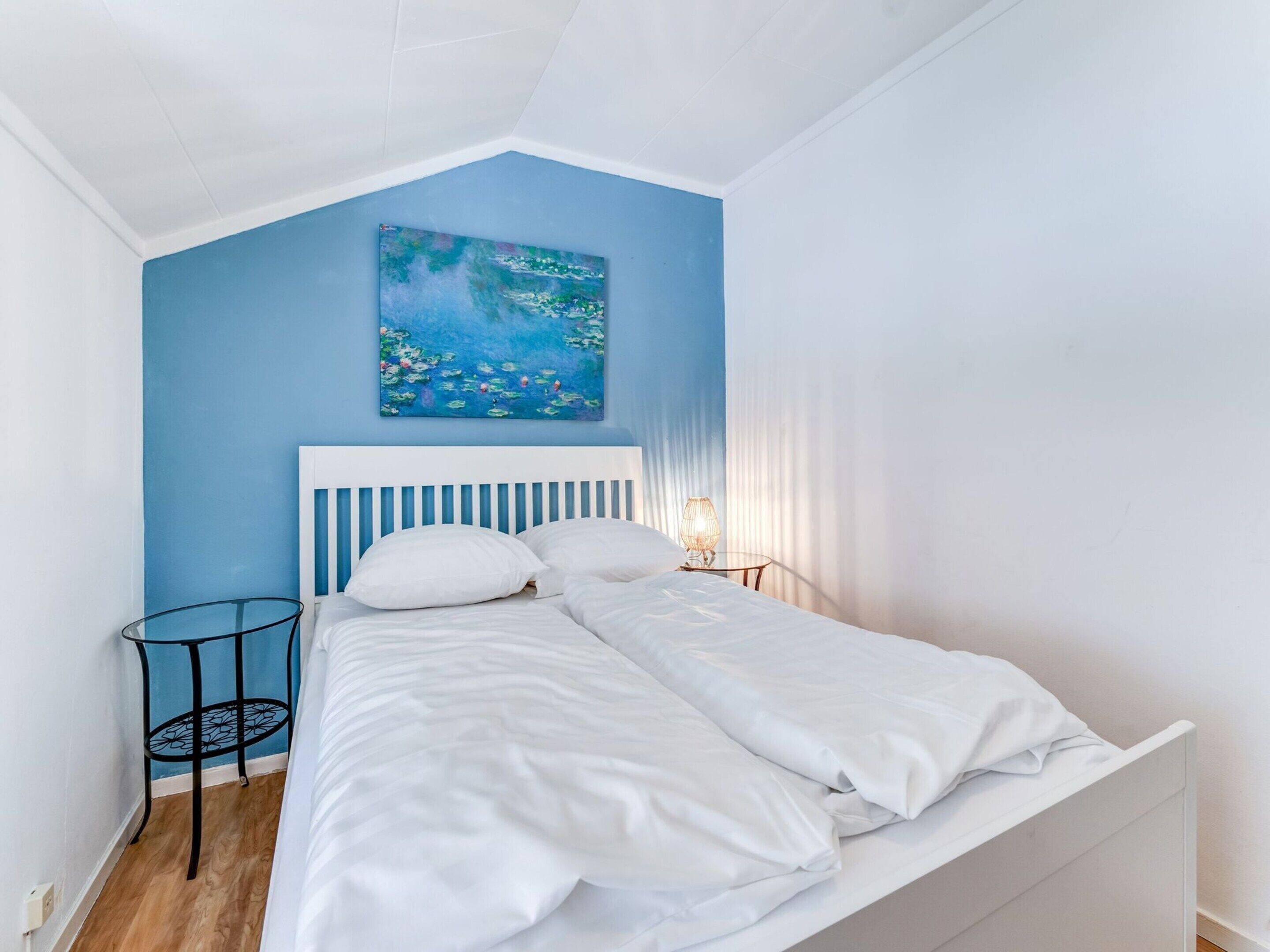 A bedroom with a white bed, a blue wall, and a painting of water lilies.