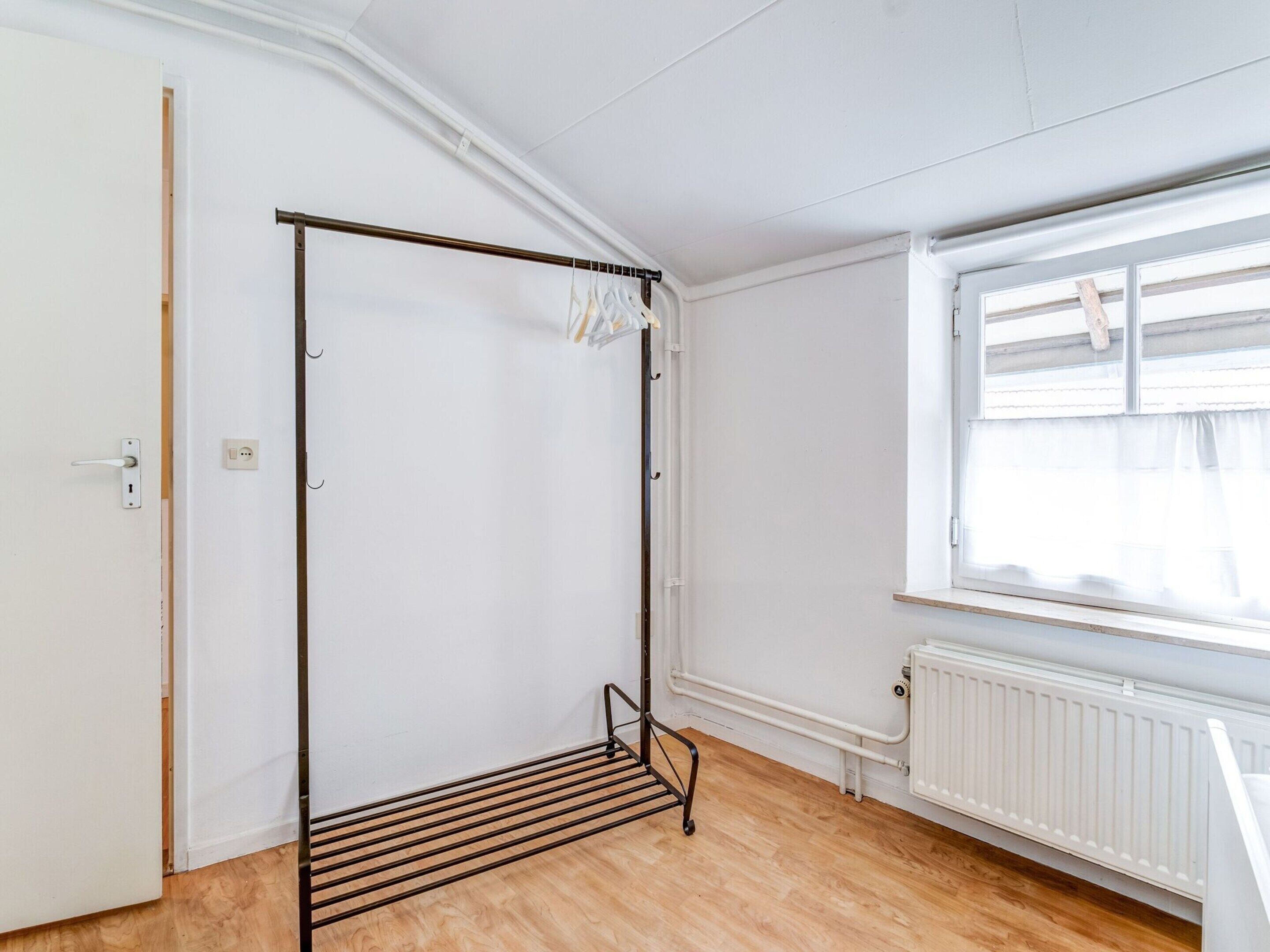 Una habitación con un tendedero de metal, un radiador, una ventana con cortina y una puerta con manija.