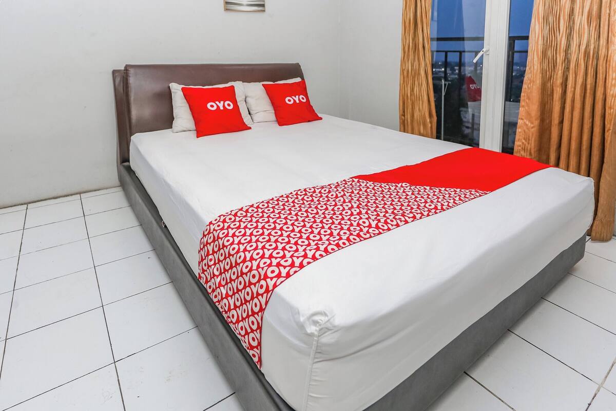 A neatly made bed with a white and red patterned bedspread, two red pillows with "OYO" text, and a view of the city through a window with curtains.
