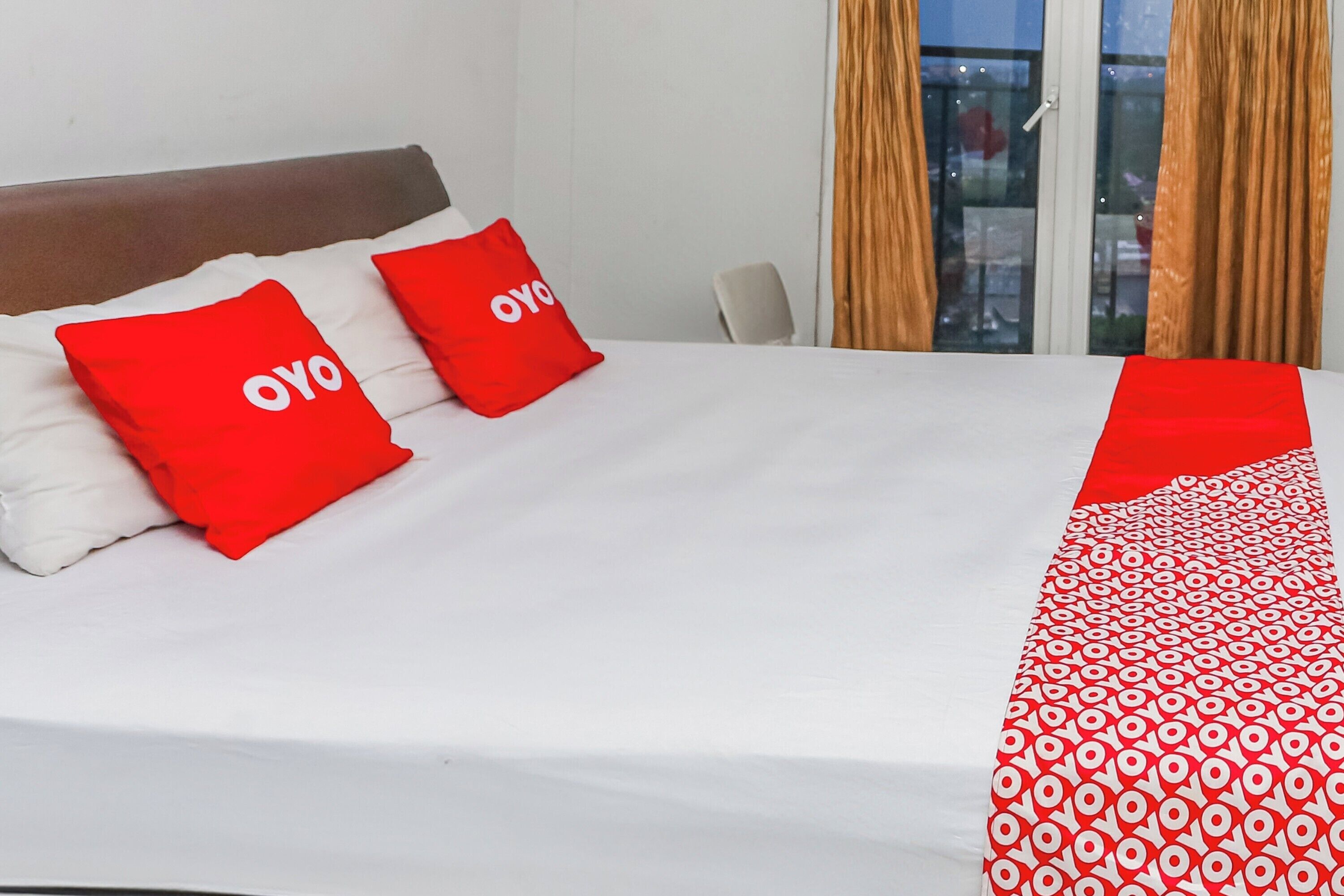 A hotel room with a bed, two red pillows with "OYO" branding, a white bedspread, and a red and white patterned throw.