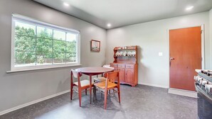 A dining area with a round table and two chairs, a wooden cabinet with dishes, a window with a view of trees, and a door. - Retro Refuge On Woodstock Dr. (Van Wert)