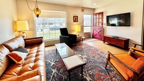 A living room with a leather sofa, a glass coffee table, a patterned rug, a wooden side table, and a television mounted on the wall. - Retro Refuge On Woodstock Dr. (Van Wert)