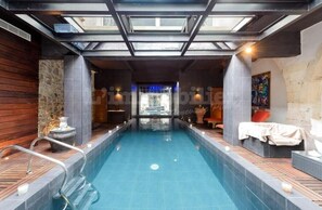 A modern indoor swimming pool with a skylight ceiling, stone walls, and a seating area with cushions. - Maison D'exception Avec Piscine Intérieure (Aigues-Mortes)
