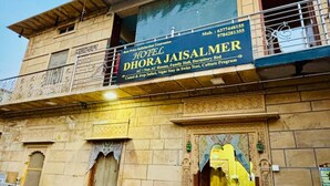 Um edifício com a placa Hotel Dhora Jaisalmer, oferecendo comodidades como quartos com e sem ar condicionado, salão familiar, beliches, safári de camelo e jipe, e programação cultural.