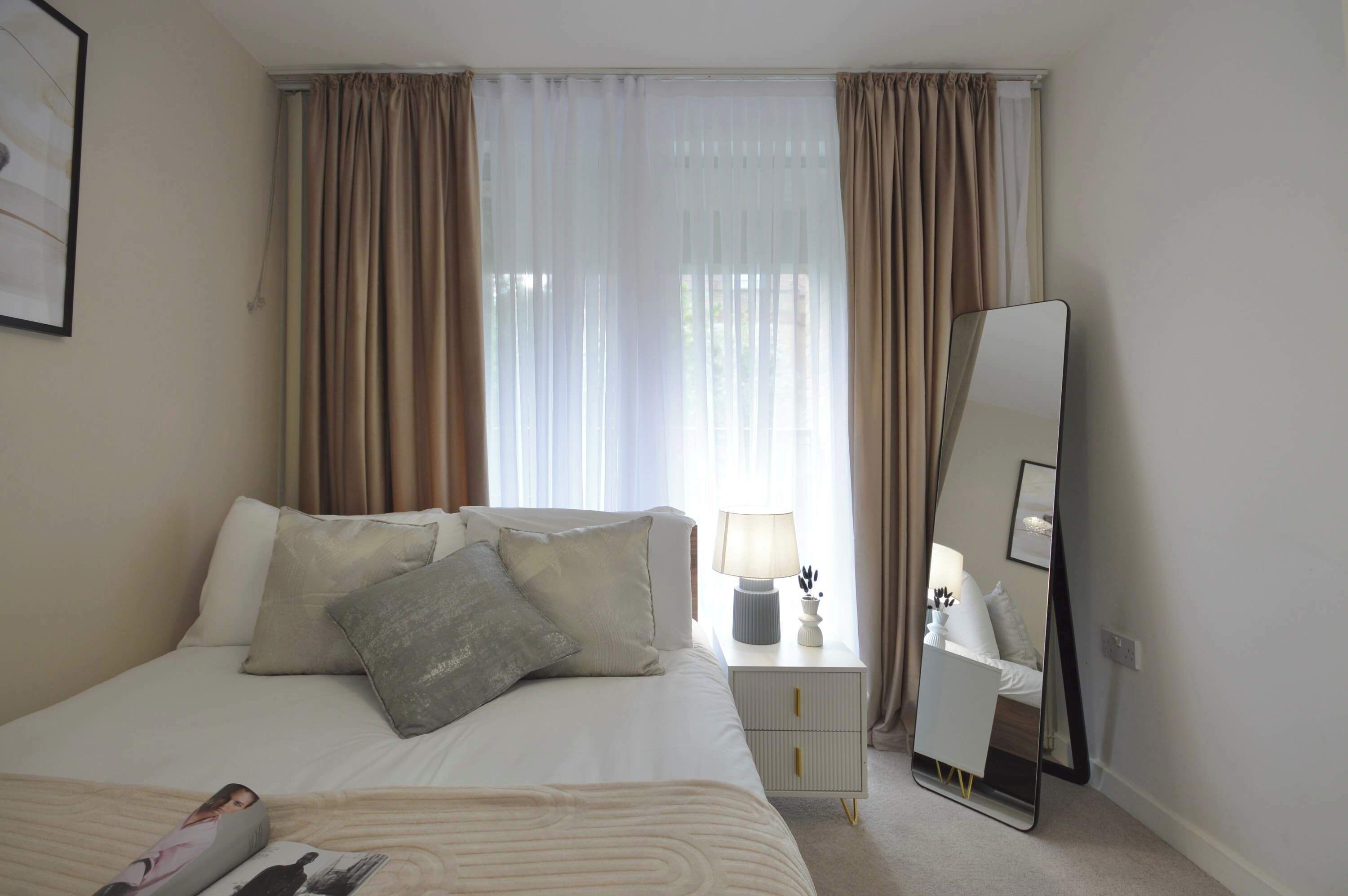 A hotel room with a bed, a nightstand with a lamp, a mirror, and a window with curtains.