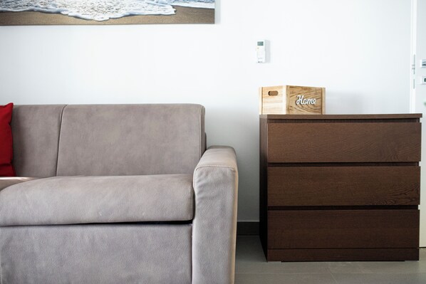 A neutral-toned sofa with a red pillow, a wooden cabinet with drawers, and a wall-mounted artwork above the sofa. - Sole & Pepe (Castellabate)