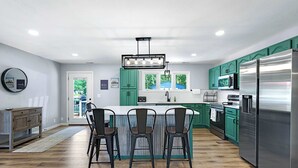 A modern kitchen with teal cabinets, a stainless steel refrigerator, and a central island with chairs. - The Lake Shack Waterfront Life (Camdenton)