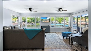A spacious living room with a large sofa, a coffee table, and a TV. There are two ceiling fans, large windows with a view of the outdoors, and a small balcony with a railing. - The Lake Shack Waterfront Life (Camdenton)