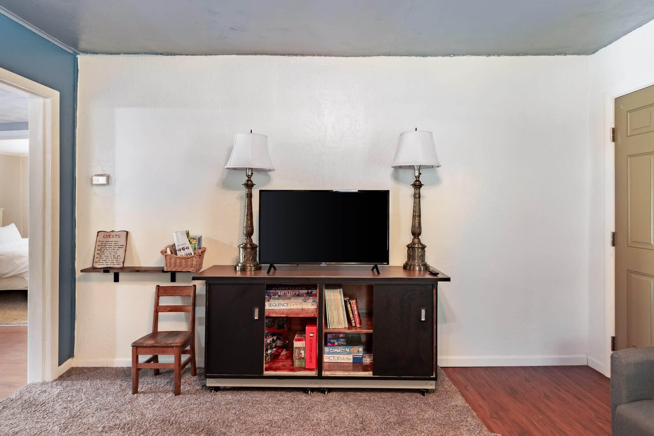 Una sala de estar con un televisor de pantalla plana, dos lámparas, una estantería y una silla de madera.