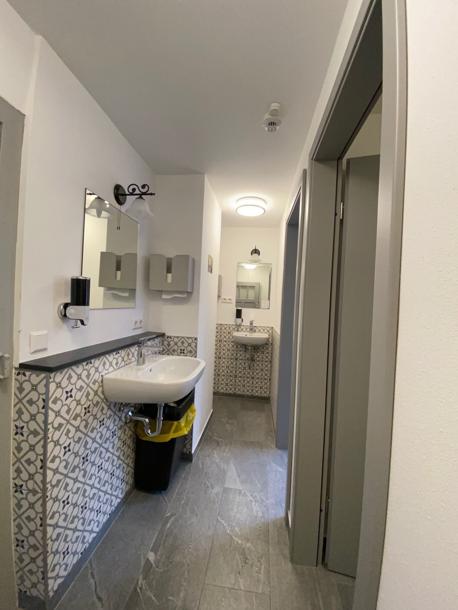 Ein schmales Badezimmer mit einem weißen Waschbecken, einem Spiegel und einer Toilette. Die Wände sind mit einem geometrischen Muster gefliest, und es steht ein Mülleimer mit gelbem Beutel dort.