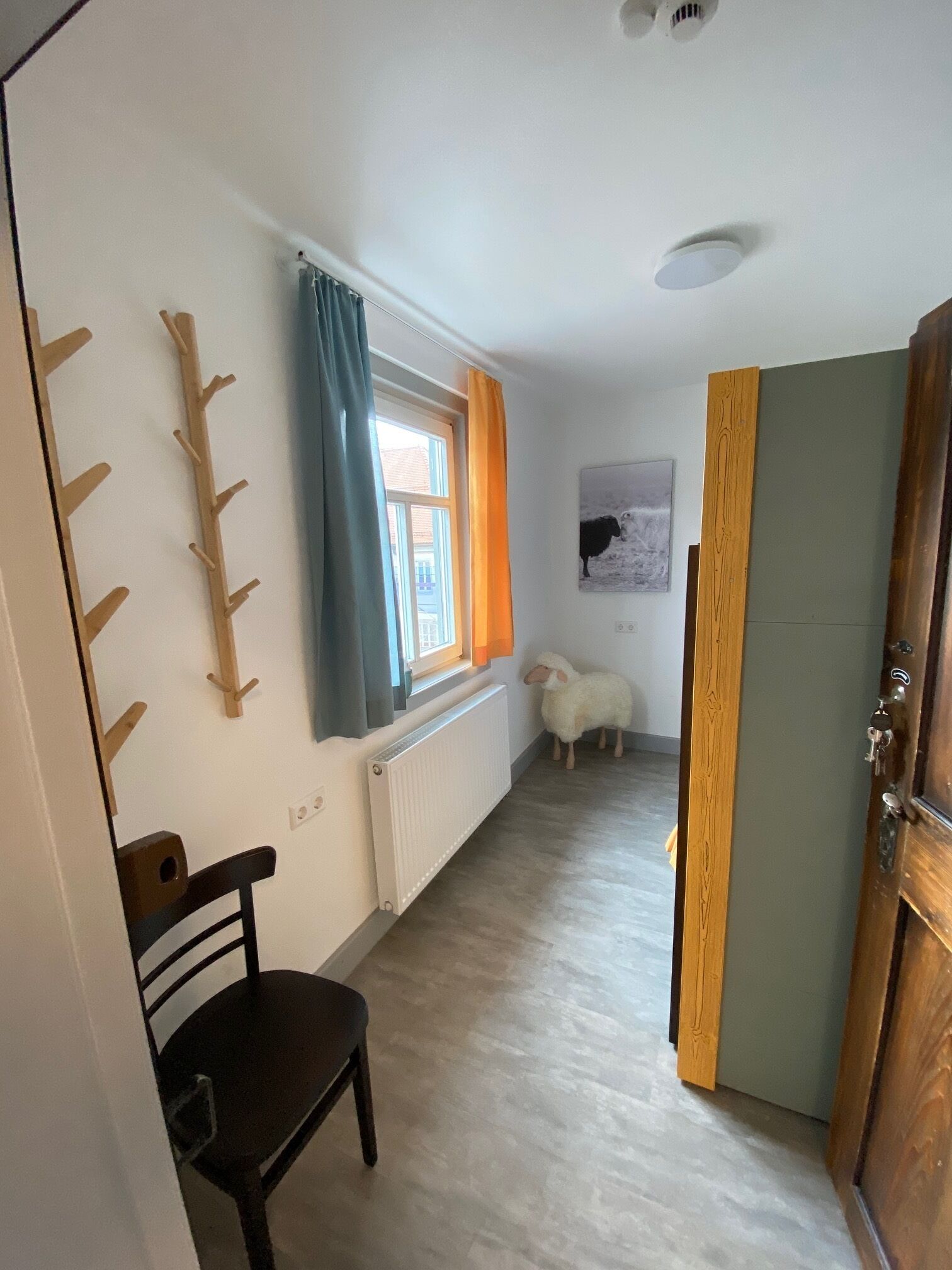 Um corredor estreito com um radiador, uma janela com cortinas, uma cadeira de madeira, uma porta de madeira e um quadro na parede.