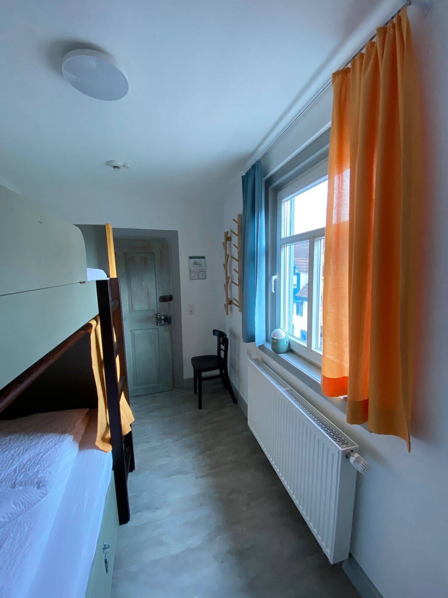 A room with bunk beds, a radiator, a window with orange curtains, and a chair.