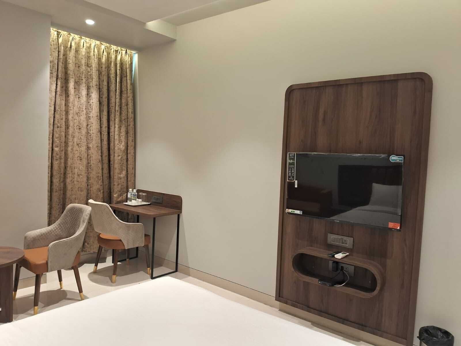 A hotel room with a desk, two chairs, a television mounted on a wooden panel, and a window with curtains.