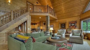 A spacious log cabin interior with a green sofa, leather armchairs, a dining area, and a wooden staircase. - Sunset Pointe | Epic Views + Hot Tub + Games (Mineral Bluff)
