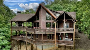A two-story wooden cabin with a balcony on the upper floor, surrounded by lush greenery. - Sunset Pointe | Epic Views + Hot Tub + Games (Mineral Bluff)