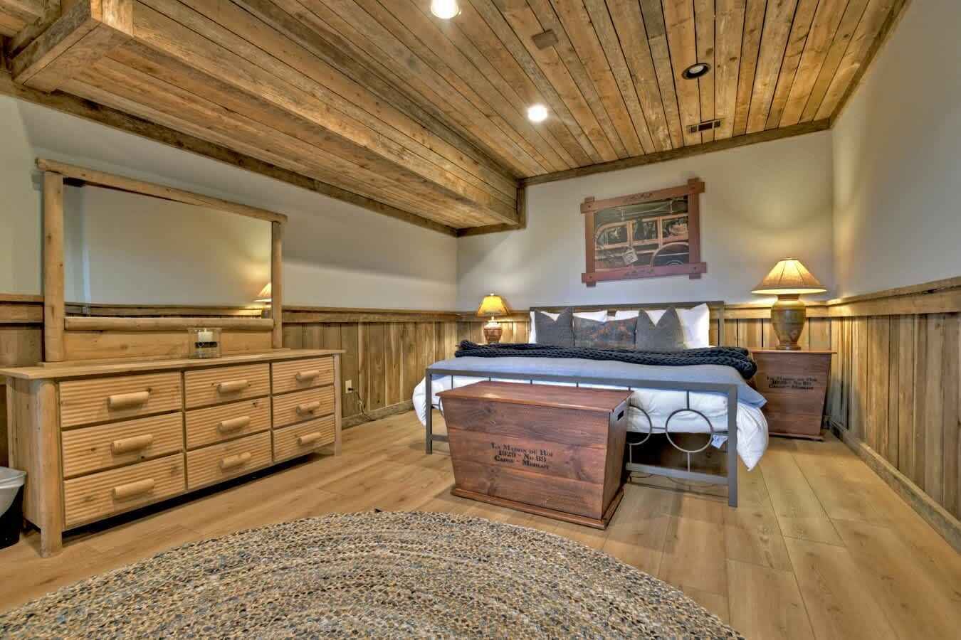 Un dormitorio con techo de madera, una cama con cabecera, un aparador, dos mesitas de noche con lámparas y una alfombra.
