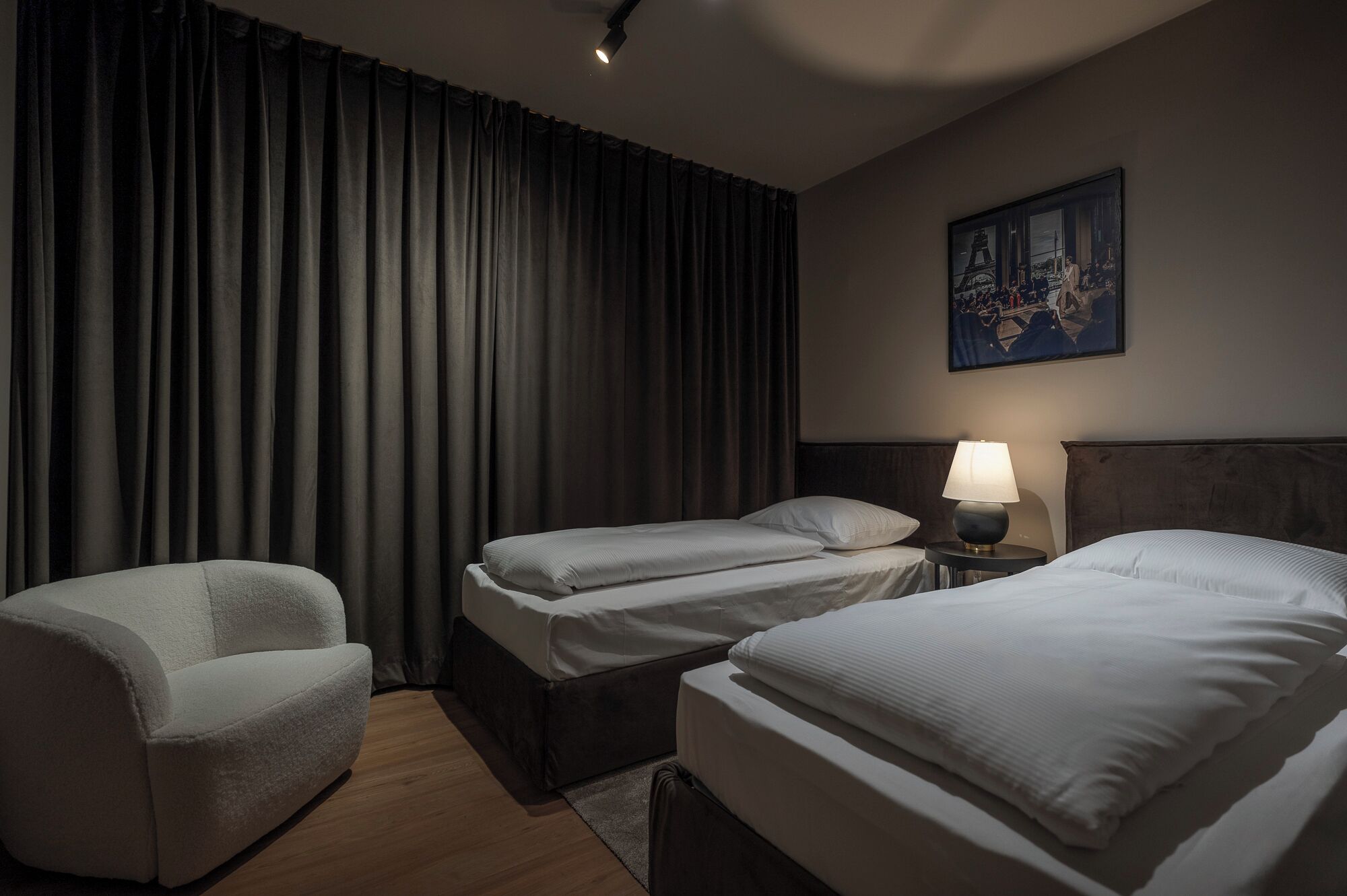 A hotel room with two single beds, a sofa, a nightstand with a lamp, and a painting on the wall.