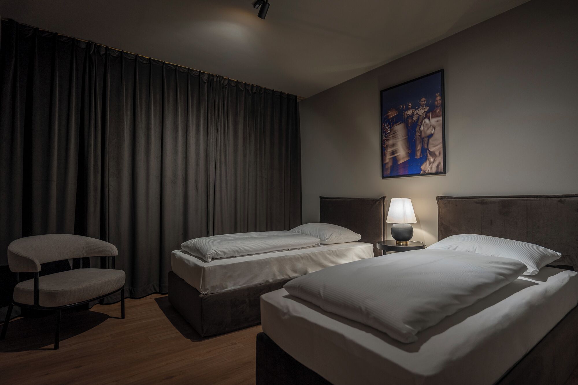 A hotel room with two beds, a chair, a lamp, and a painting on the wall.