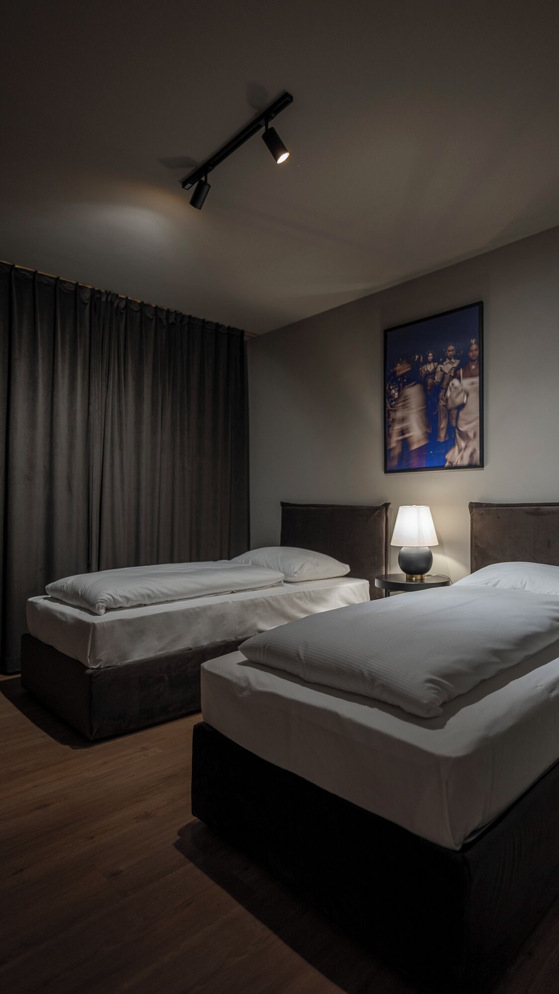 Ein Hotelzimmer mit zwei Betten, einem dunklen Vorhang, einem Gemälde an der Wand und einer Lampe auf einem Nachttisch.